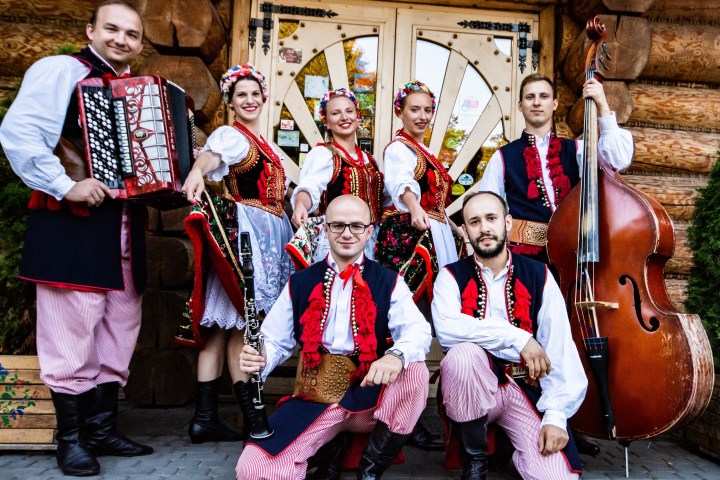 Group in traditional costumes with musical instruments, posing in front of a wooden building.