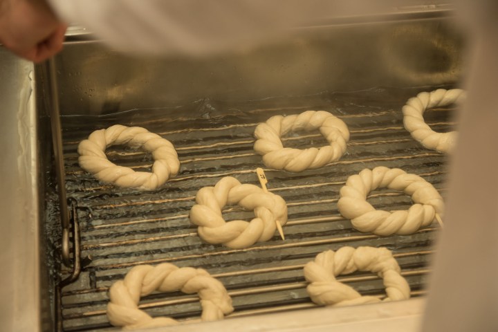 Dough rings boiling on a metal rack in hot water, with a hand holding a tool.