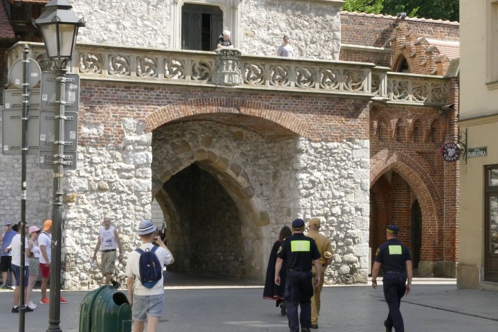 People walking near an old brick and stone archway with a balcony above.