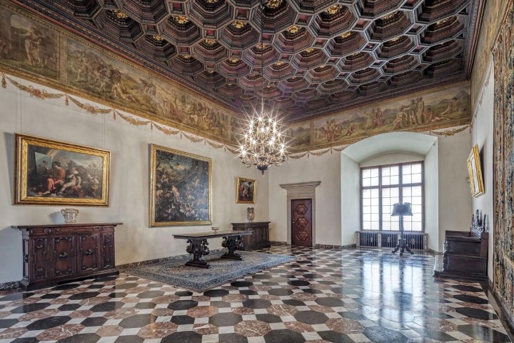 Ornate room with patterned ceiling, chandelier, paintings, and checkerboard floor.