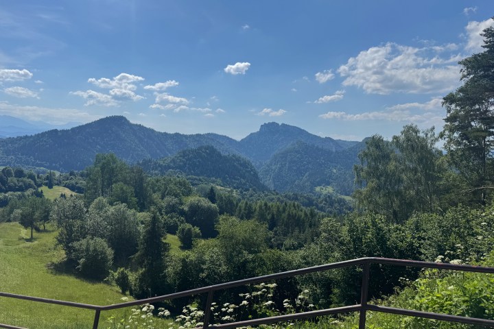 Mountainous landscape with trees and a metal railing under a blue sky with clouds.