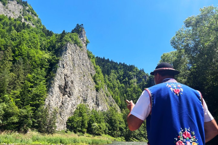 Man in traditional attire beside river and rocky hill with trees.