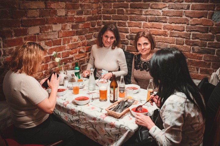 Four women dining at a table with drinks and bowls of soup, brick wall backdrop.