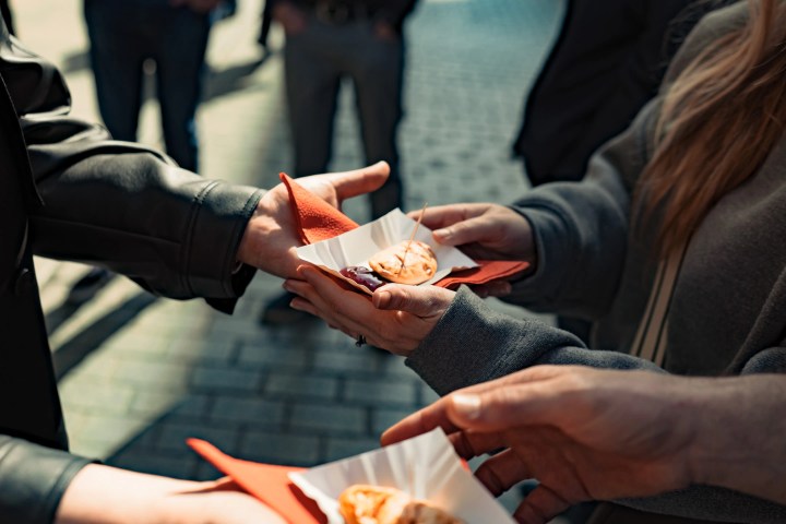 Hands exchanging pastries on paper trays in an outdoor setting.