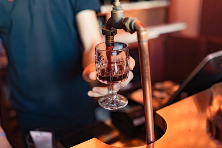 Person filling a glass with red wine from a tap.