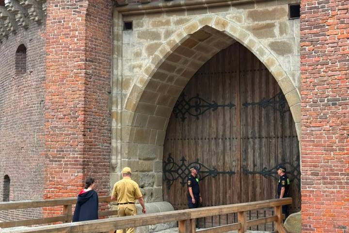People stand outside a large, arched wooden gate in a red brick building.