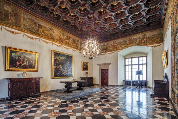 Opulent room with chandeliers, ornate ceiling, paintings, and patterned floor tiles.
