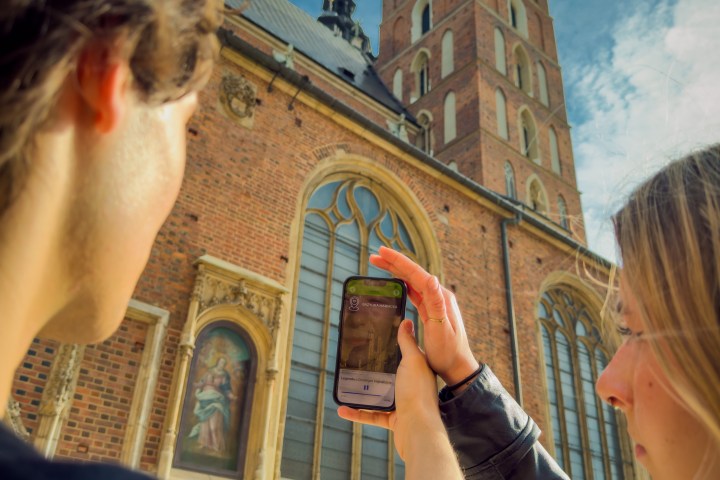 Two people taking a photo of a brick church with a tall tower.