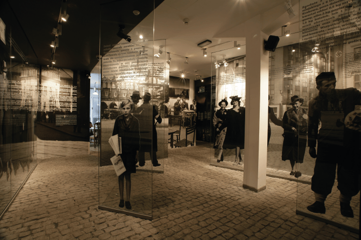 Museum exhibit with historical photos on glass panels and cobblestone floor.