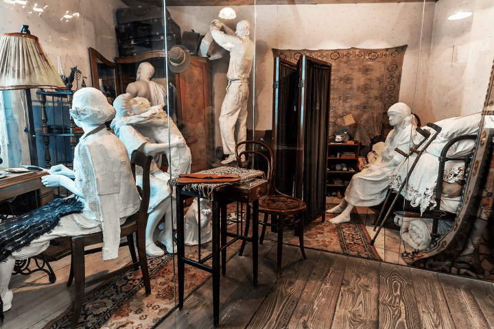 Room with mannequin figures, vintage furniture, ornate rugs, and a small bed.
