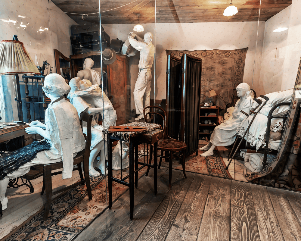 Room with mannequin figures, vintage furniture, ornate rugs, and a small bed.