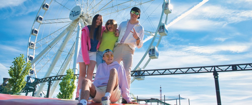 Group of four people posing in front of a Ferris wheel on a sunny day.