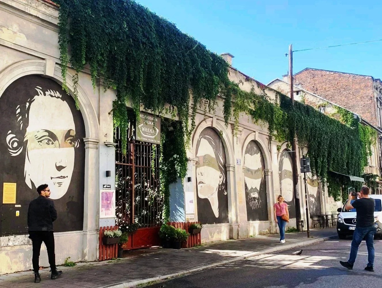 Street with wall murals of faces, greenery cascading down arches, people walking nearby.