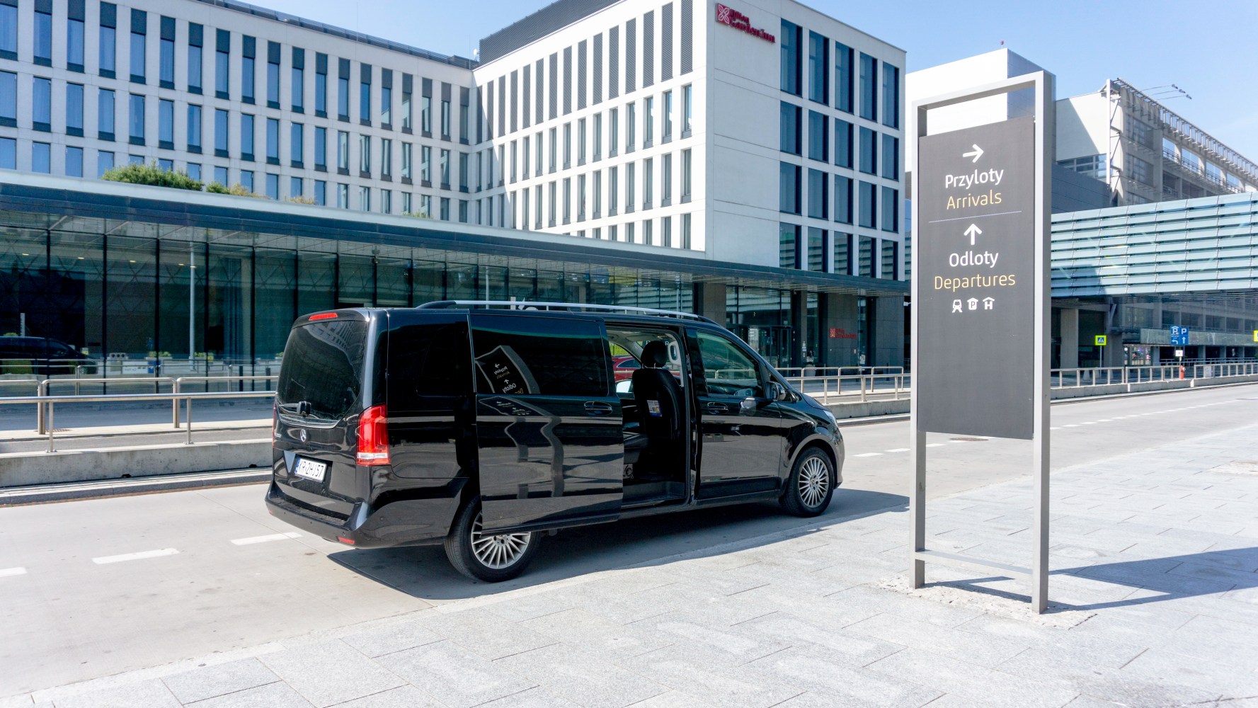 Black van parked outside a modern airport building next to an arrivals and departures sign.