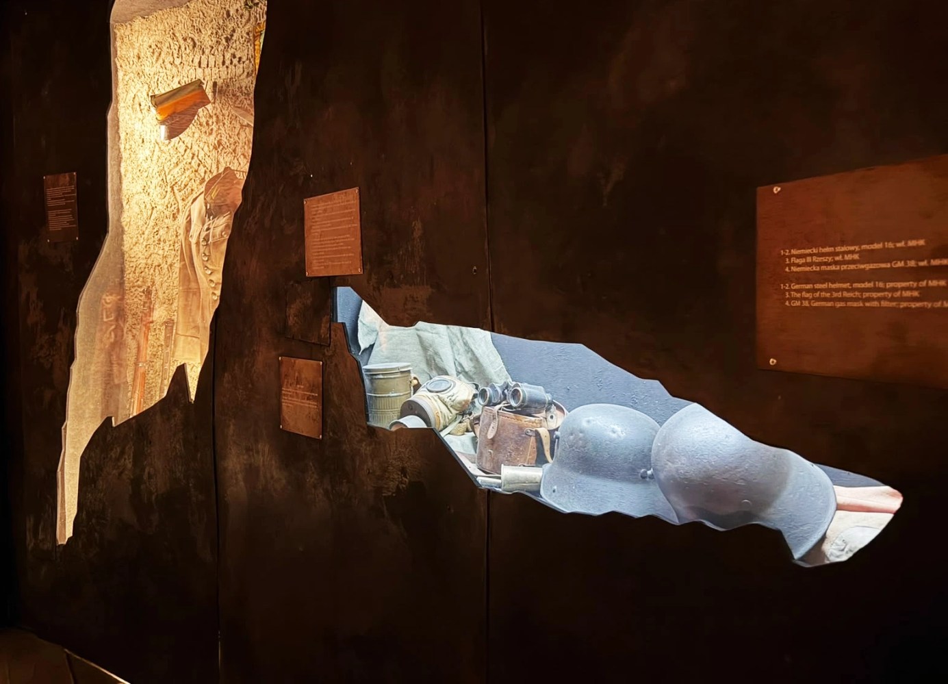 Museum display with helmets and wartime artifacts behind uneven, jagged openings in a dark wall.
